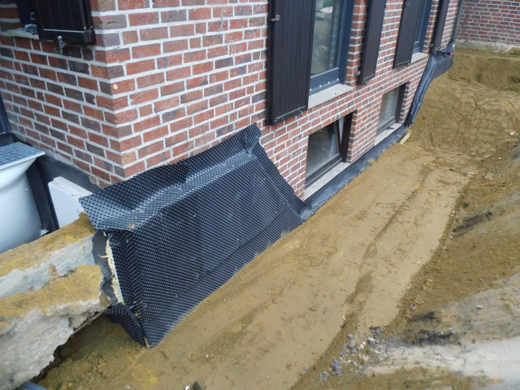 Schwarze Noppenbahn als Schutzschicht am freigelegten Sockel eines Backsteinhauses, davor eine offene Baugrube.