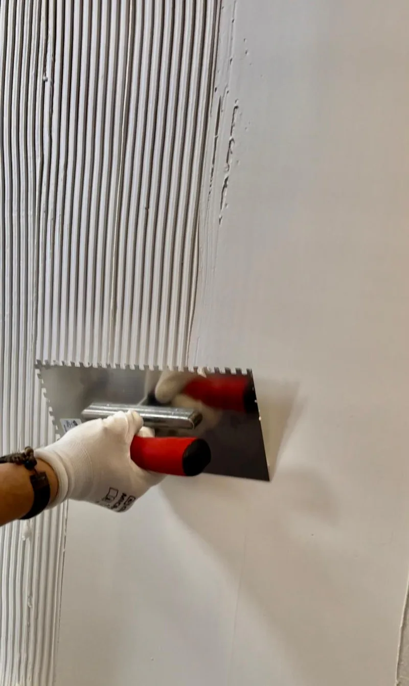 Eine behandschuhte Hand zieht eine weiße Beschichtung mit einer Zahnkelle an einer Wand auf, sodass gleichmäßige Rillen entstehen.