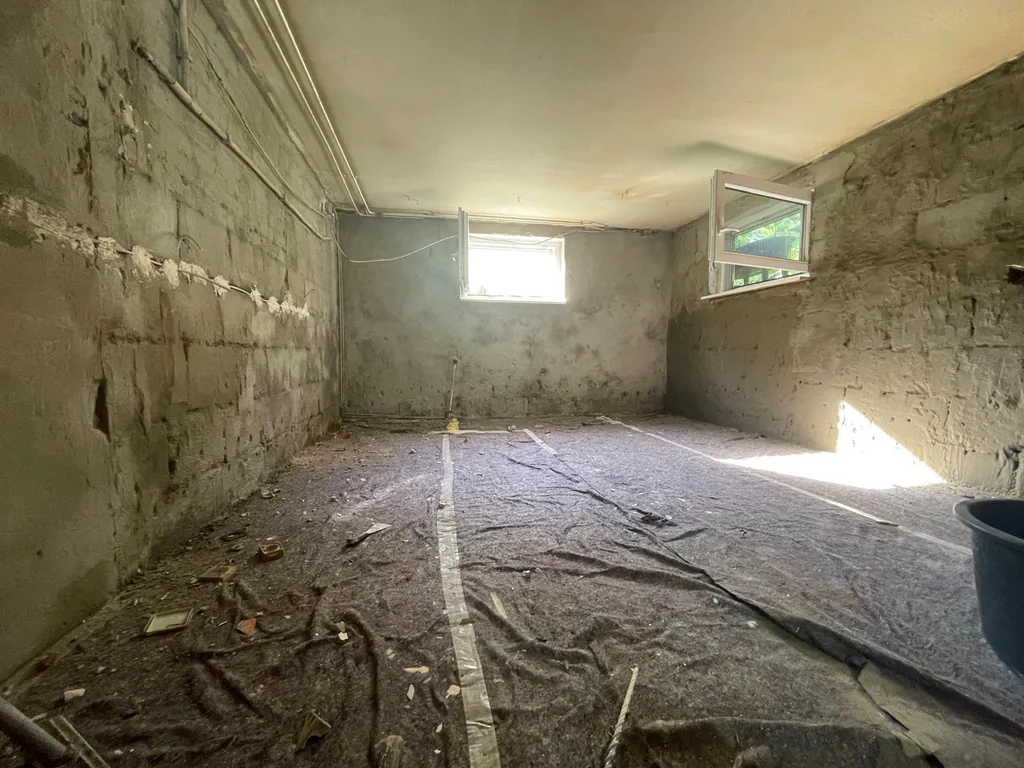 Kellerraum mit unverputztem Mauerwerk, einem waagerechten helleren Streifen auf halber Wandhöhe, abgelegter Schutzfolie und Bauschutt auf dem Boden.