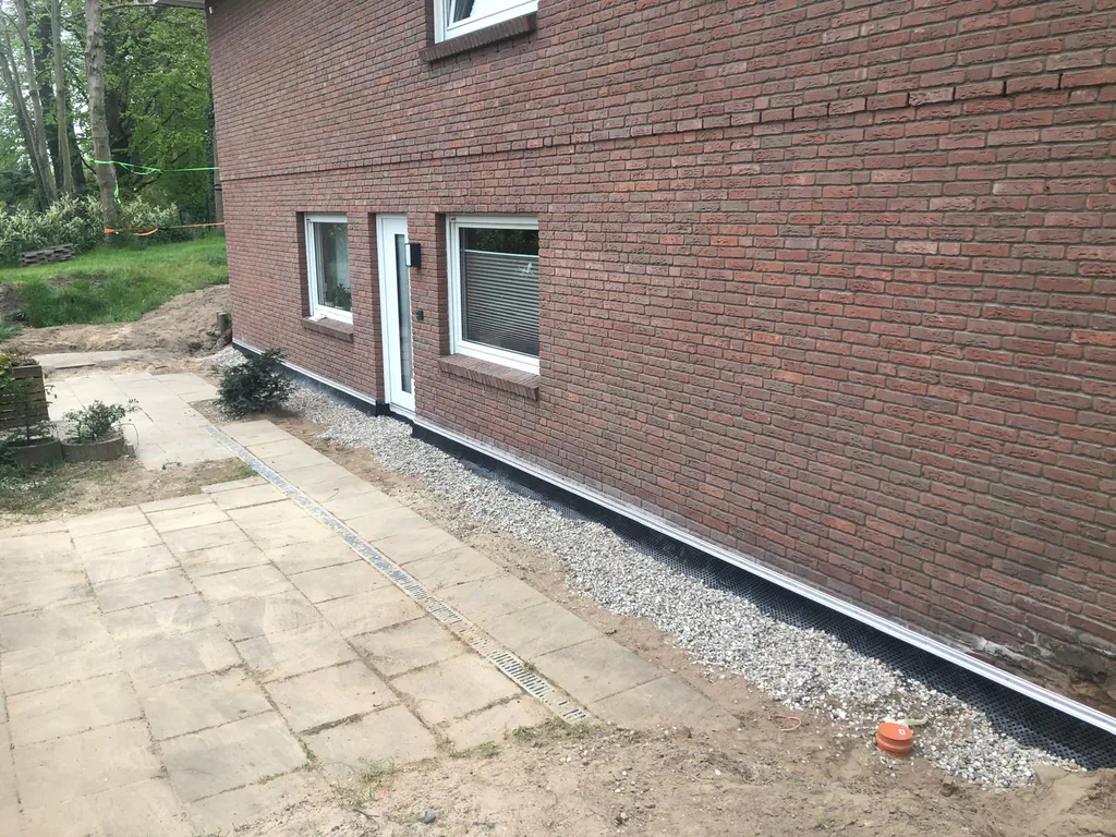 Fertige Außenanlage an einem Backsteinhaus: gepflasterter Weg, ein Kiesstreifen entlang des Sockels und eine dunkle Sockelabdichtung.