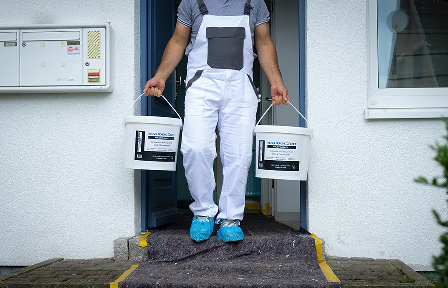Eine Person in weißer Arbeitskleidung und blauen Überschuhen trägt zwei Materialeimer über eine mit Schutzvlies ausgelegte Haustürschwelle.